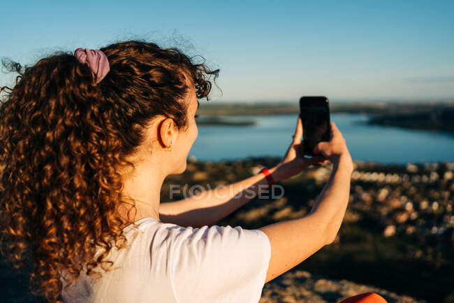 Visão traseira de caminhante feminino irreconhecível com cabelo encaracolado em roupas casuais tomando selfie sentado em cobertor em penhasco rochoso acima do mar no dia ensolarado — Fotografia de Stock