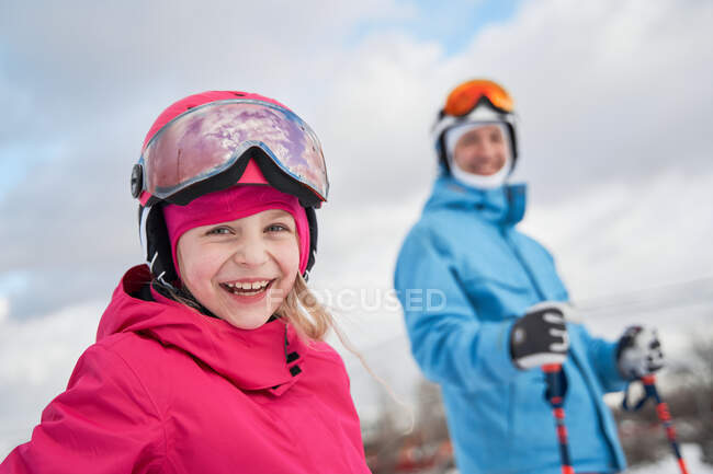 Menina alegre vestindo capacete de esqui e activewear de pé perto pai borrado em terreno de inverno nevado e olhando para a câmera com rosto sorridente — Fotografia de Stock