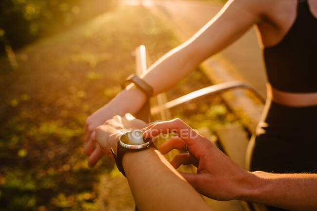 Crop unrecognizable female athlete checking pulse on smart watch while standing in park with anonymous athlete at sunset — Stock Photo