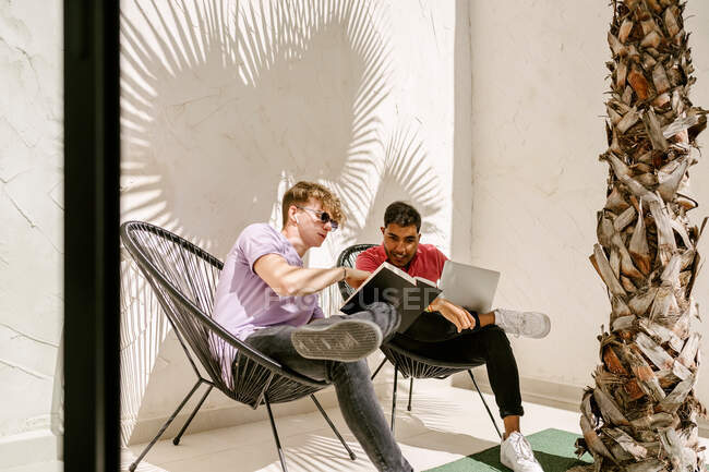 Стильні молоді багаторасові друзі-чоловіки використовують ноутбук і читають книгу, відпочиваючи на стільцях на терасі в сонячний день — стокове фото