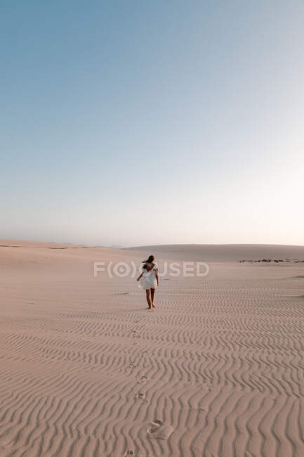 Visão traseira de turista feminino anônimo em vestido branco passeando na areia com nervuras sob o céu claro — Fotografia de Stock
