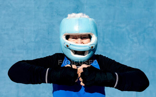 Content mature female in activewear boxing helmet and hand wraps raising hands near head against blue wall and looking at camera — Stock Photo