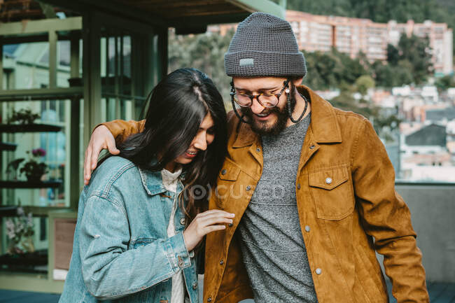Молодая стильная пара нежно обнимается, стоя на деревянной террасе рядом с домом — стоковое фото