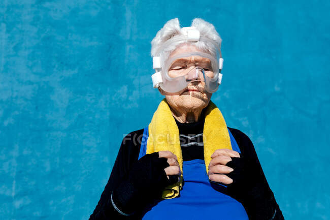Serious mature female in activewear in protective boxing head guard with towel on shoulder standing on blue background with closed eyes — Stock Photo
