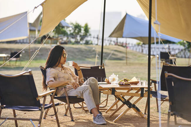 Hermosa mujer asiática étnica en gafas de sol sentada en la mesa bebiendo té mientras pasa un tiempo relajante en la zona de acampada durante las vacaciones - foto de stock