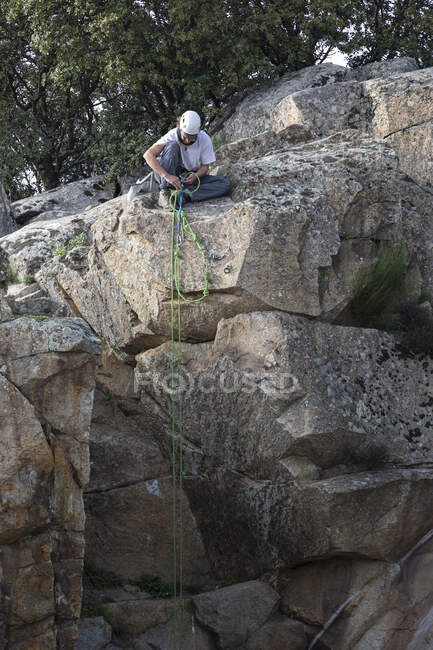 Male climber in protective helmet sitting on rock and fastening harness before descent from mountain — Stock Photo