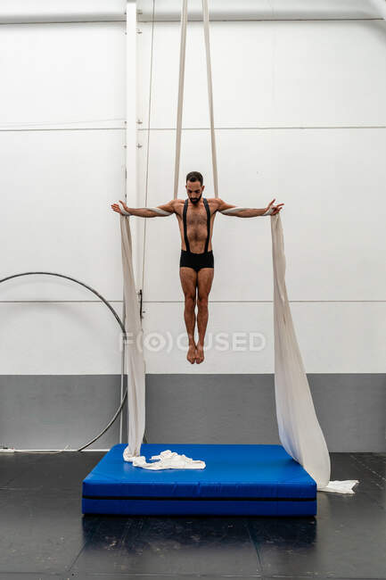 Full body muscular strong sportsman in shorts performing exercise on aerial silks in modern light fitness center — Stock Photo