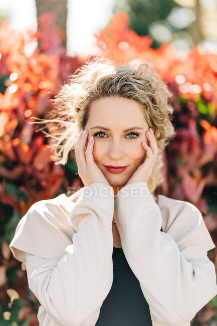 Mujer rubia feliz en vestido elegante y abrigo de pie tocando la cara mientras está entre los árboles mirando a la cámara - foto de stock