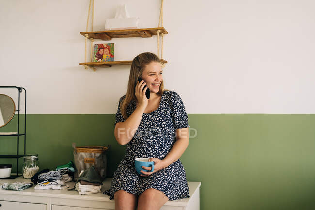 Fröhliche, erwartungsvolle Frau mit einer Tasse Heißgetränk spricht auf dem Handy, während sie im Hauszimmer wegschaut — Stockfoto