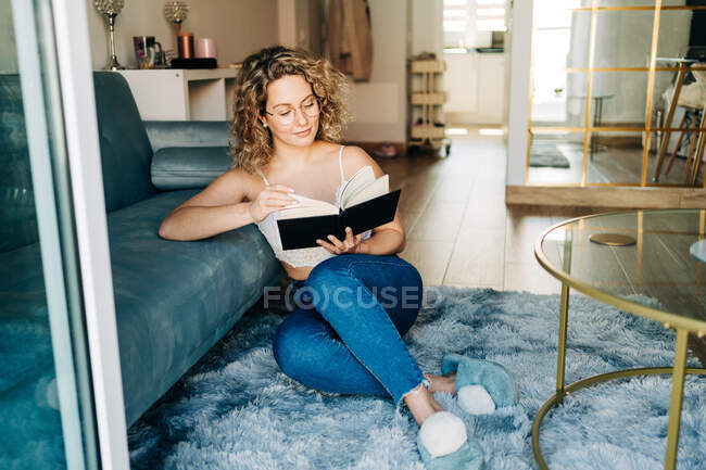 Полное тело позитивной молодой женщины с кудрявыми волосами в повседневной одежде и тапочках, сидящих на ковре возле дивана и читающих интересную книгу в выходные дни дома — стоковое фото