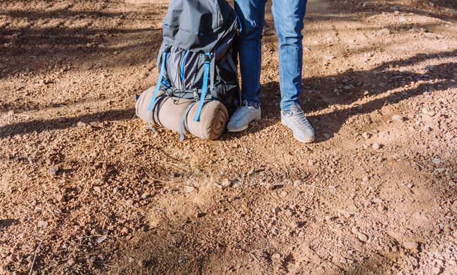 Анонимный турист в джинсах и кроссовках, стоящий на грунтовой дороге возле большого рюкзака в солнечный день — стоковое фото