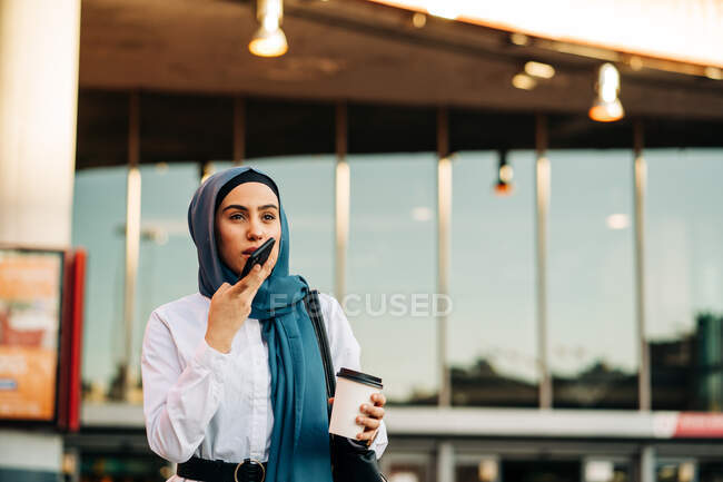 Ethnic female in headscarf standing with takeaway drink on street and recording voice message on mobile phone — Stock Photo