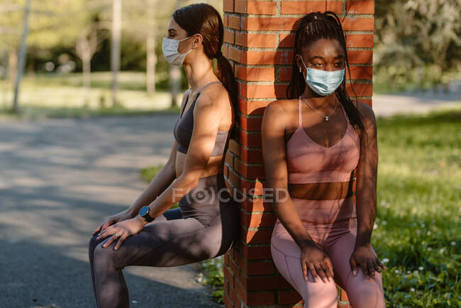 Atletas femeninas multirraciales con máscaras desechables en cuclillas contra postes ásperos mientras miran hacia otro lado durante el entrenamiento en el parque urbano - foto de stock