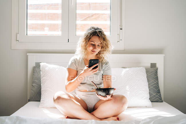 Frohe junge Frau in häuslicher Kleidung, die Fotos von leckeren Frühstück in Schüssel macht, während sie morgens mit gekreuzten Beinen auf dem gemütlichen Bett sitzt — Stockfoto