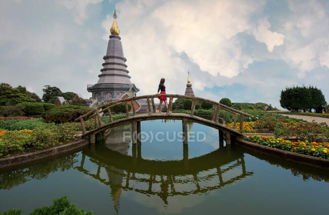 Seitenansicht einer anonymen Touristin, die auf einer Brücke über einen Teich spaziert und dabei einen alternden orientalischen Schrein in Thailand betrachtet — Stockfoto