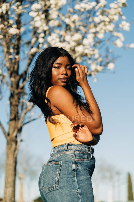 Low angle side view of beautiful African American female standing in blooming spring park and enjoying sunny weather looking at camera — Stock Photo