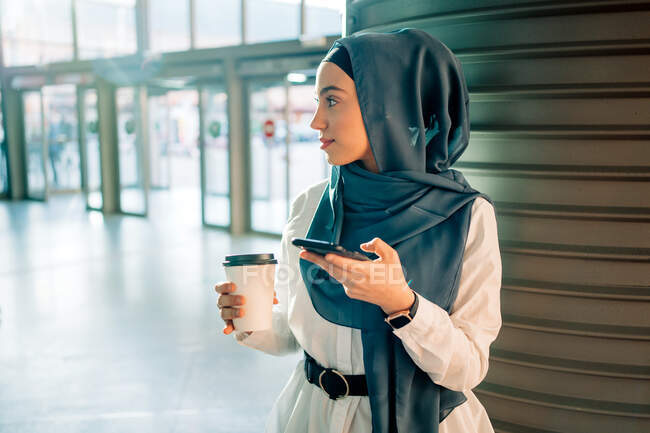 Donna etnica in hijab in piedi sulla stazione e la navigazione cellulare in attesa di treno — Foto stock