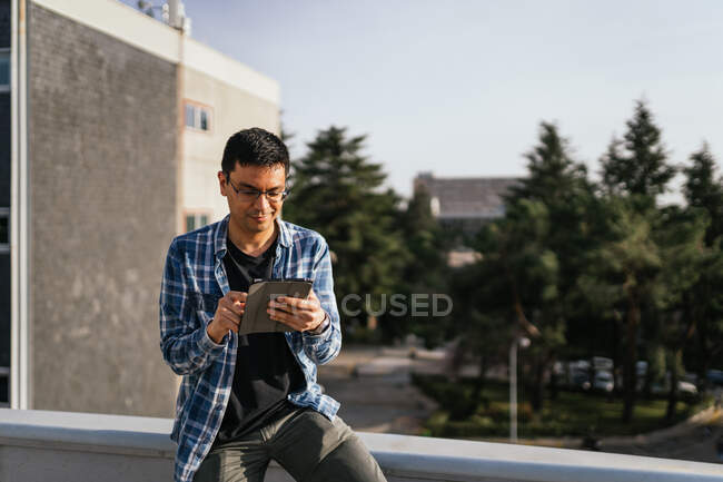 Концентрированный мужчина в очках и клетчатой рубашке, просматривающий современные планшеты, стоя на крыше в пригороде в солнечный летний день — стоковое фото