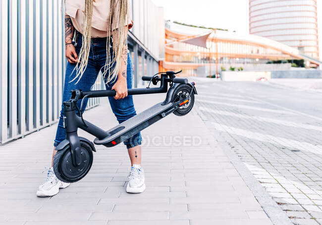 Crop anonimo femminile in abbigliamento casual in piedi con scooter moderno sulla passerella piastrellata in città — Foto stock