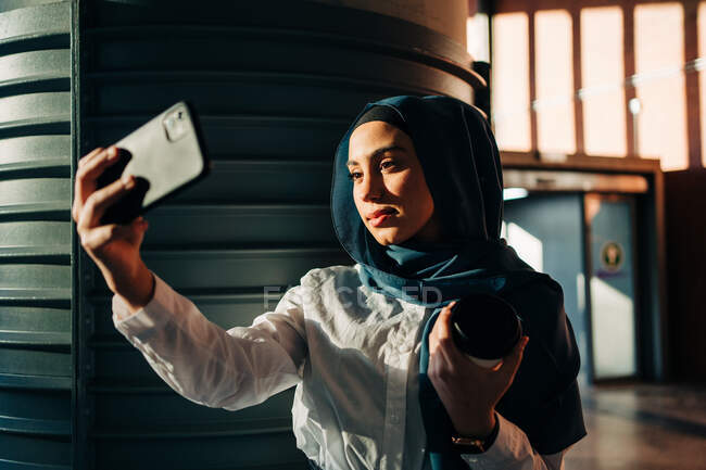 Femme ethnique en hijab debout sur le quai de la gare et prenant selfie sur téléphone portable en attendant le train — Photo de stock