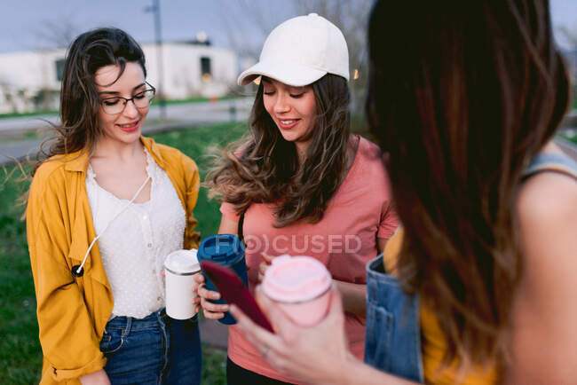 Crop content best female friends with hot beverages in tumblers speaking while looking at each other in city — Stock Photo