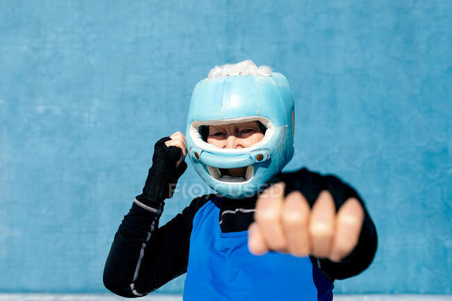 Вміст зрілої жінки в шоломі боксу та обгортаннях рук, що піднімають руки біля голови на синю стіну та дивляться на камеру — стокове фото