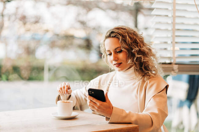 Conteúdo fêmea sentada à mesa na cafeteria e mexendo bebida saborosa na xícara enquanto navega no smartphone — Fotografia de Stock