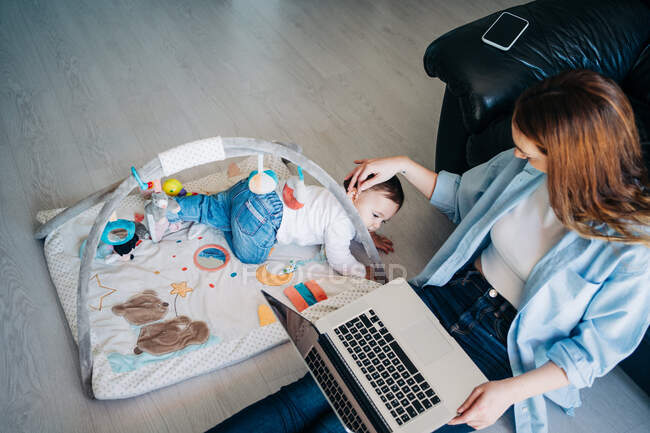 Top view of mother wearing casual outfit sitting on floor with netbook on laps and outstretching hand to cute little baby playing with toys on floor — Stock Photo