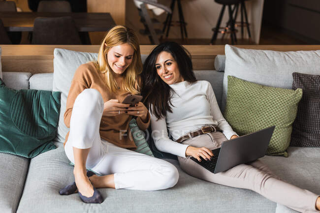 Paar fröhlicher homosexueller Frauen, die auf der Couch chillen und Videos auf dem Smartphone ansehen und am Laptop arbeiten, während sie am Wochenende zusammen unterhalten — Stockfoto