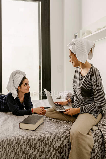 Happy young ethnic lady in casual clothes and towel on head lying on bed and looking at girlfriend working remotely on laptop at home — Photo de stock