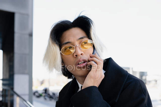 Молодой внимательный азиатский мужчина в формальной одежде и солнцезащитных очках по телефону на смартфоне при дневном свете — стоковое фото