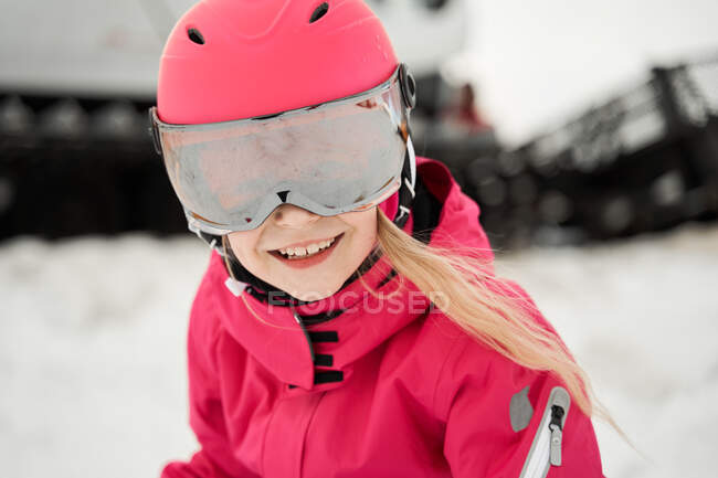 Positive cute girl in pink warm activewear goggles and helmet skiing alongside snowy slope on clear winter day — Stock Photo