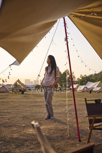 Étnica mujer asiática tener un tiempo relajante de pie mirando hacia otro lado en el área de camping durante la puesta del sol - foto de stock
