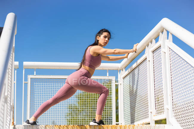 Low angle side view of fit young female athlete in sportswear leaning on fence during training and looking away — Fotografia de Stock