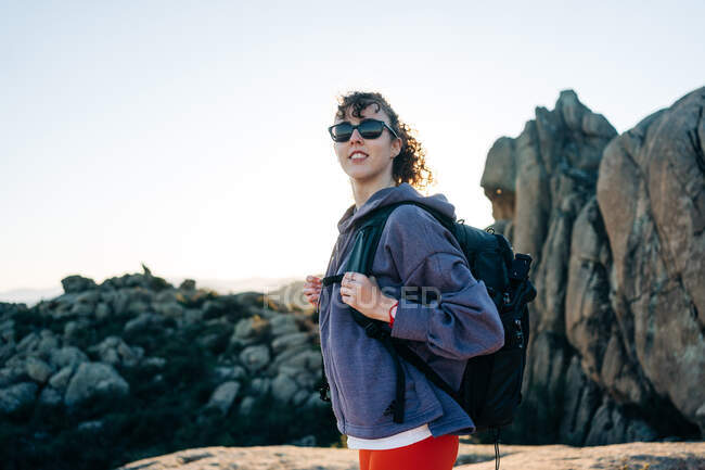 Vista lateral de la joven exploradora alegre en sudadera con capucha y gafas de sol sonriendo mientras admira la naturaleza durante el trekking en el valle montañoso rocoso en el día soleado - foto de stock