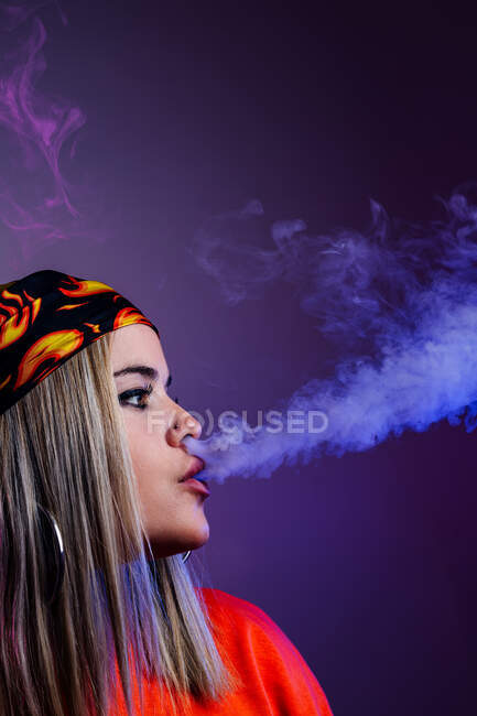 Vista lateral de la hembra fresca en traje de calle fumar cigarrillo electrónico y la exhalación de humo a través de la nariz en el fondo púrpura en el estudio con iluminación de neón rosa - foto de stock