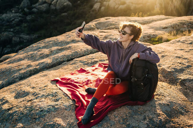 Радостная молодая туристка с вьющимися волосами в повседневной одежде и солнцезащитных очках, опираясь на рюкзак и делая селфи со смартфоном, расслабляясь на склоне скалистого холма в солнечный день — стоковое фото