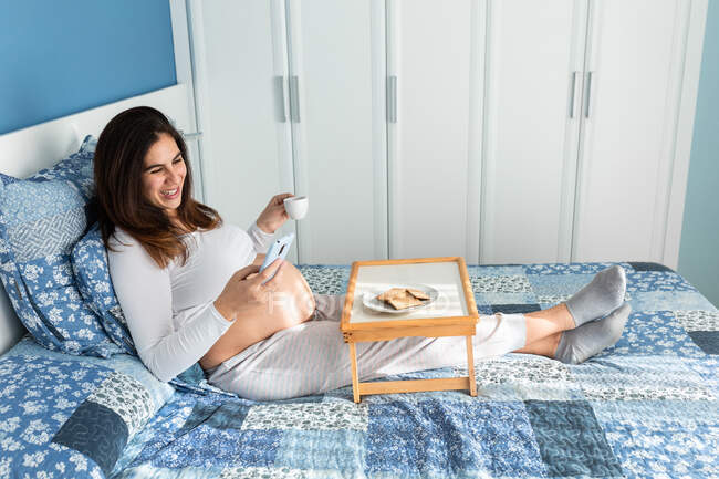 Vue latérale de gaie femme enceinte assise sur le lit le matin et prenant le petit déjeuner tout en naviguant sur smartphone — Photo de stock