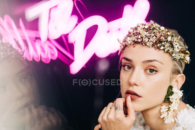 Charming tender young bride in white lace gown and luxurious floral wreath and earrings looking at camera against black background with neon lights — Photo de stock