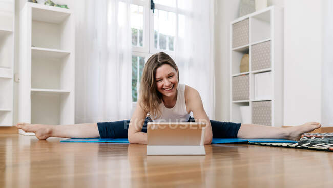 Lächeln flexible Frau praktiziert Yoga in Upavista Konasana, während sie auf Matte sitzt und Online-Video-Tutorial auf dem Tablet zu Hause anschaut — Stockfoto