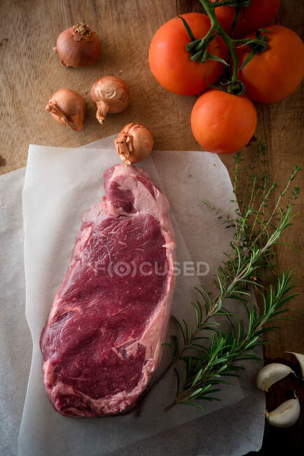 Overhead view of uncooked meat piece against ripe tomatoes and unpeeled onions with garlic cloves on brown background — Stock Photo