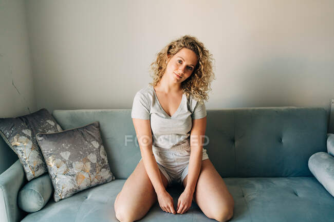 Full length positive attractive female in shorts sitting on knees on cozy couch in living room and looking at camera — Foto stock