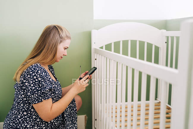 Vista laterale della giovane donna incinta che chatta sul cellulare contro la culla nella stanza della casa — Foto stock