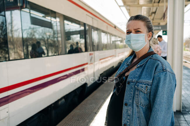 Молодая женщина в повседневной одежде и защитной маске стоит на железнодорожном вокзале и смотрит в сторону, ожидая поезда — стоковое фото