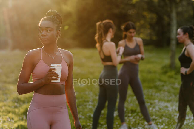 Atleta afroamericana en ropa deportiva de pie con taza de bebida en el parque por la noche y mirando hacia otro lado - foto de stock