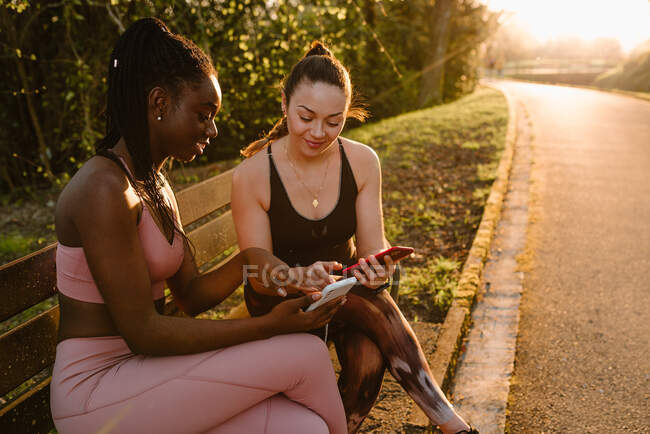 Весела багаторасова жінка-спортсменка в активному одязі сидить на лавці в парку і використовує мобільні телефони разом після тренування на заході сонця — стокове фото