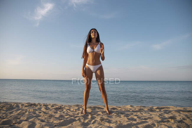 Young barefoot female in swimwear looking at camera while standing on sandy shore against ocean under cloudy blue sky — Stock Photo