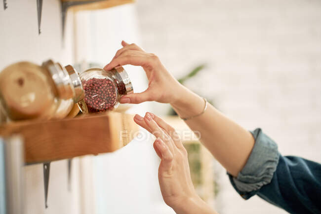 Crop unrecognizable female taking glass jar of red peppercorns from wooden shelf with assorted spices in house — Stock Photo