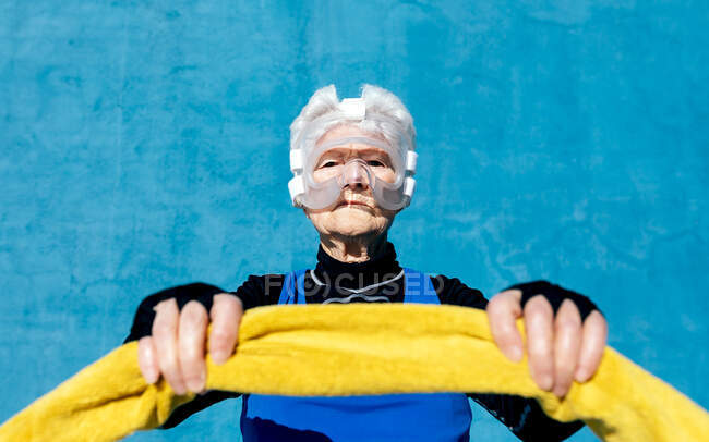 Grave maduro fêmea em activewear no protetor de cabeça de boxe de proteção com toalha nas mãos em pé sobre fundo azul e olhando para a câmera — Fotografia de Stock
