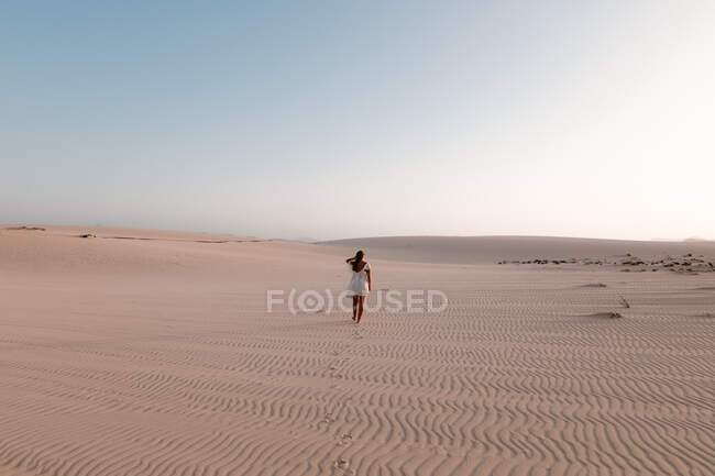 Back view of anonymous female tourist in white dress strolling on ribbed sand under light sky — Stock Photo